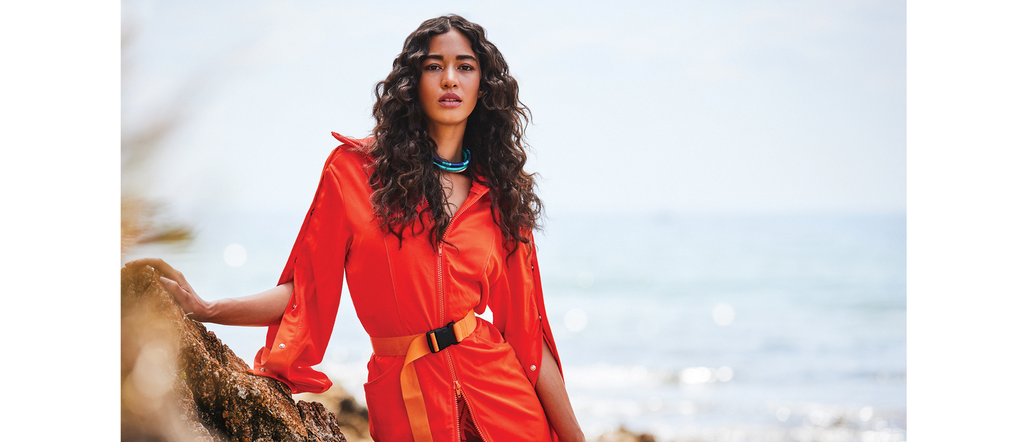 Woman with naturally textured, curly hair in a vibrant red outfit standing on rocky shoreline, demonstrating an effortless and striking hairstyle.